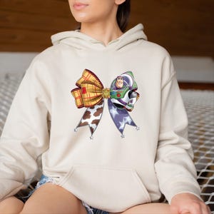 May include: A beige hoodie with a vibrant bow design. The bow incorporates a plaid pattern, a cow print, and a section with a purple star pattern. The design also includes a graphic of Buzz Lightyear.