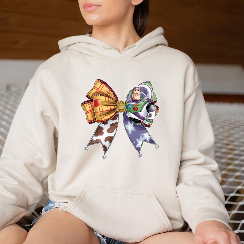 May include: A beige hoodie with a vibrant bow design. The bow incorporates a plaid pattern, a cow print, and a section with a purple star pattern. The design also includes a graphic of Buzz Lightyear.