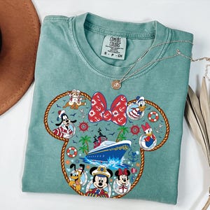 May include: A seafoam green t-shirt with a Minnie Mouse head design incorporating nautical themes. The design includes a red bow with white anchors, a cruise ship, and Disney characters in sailor outfits. A silver necklace is placed on the shirt.