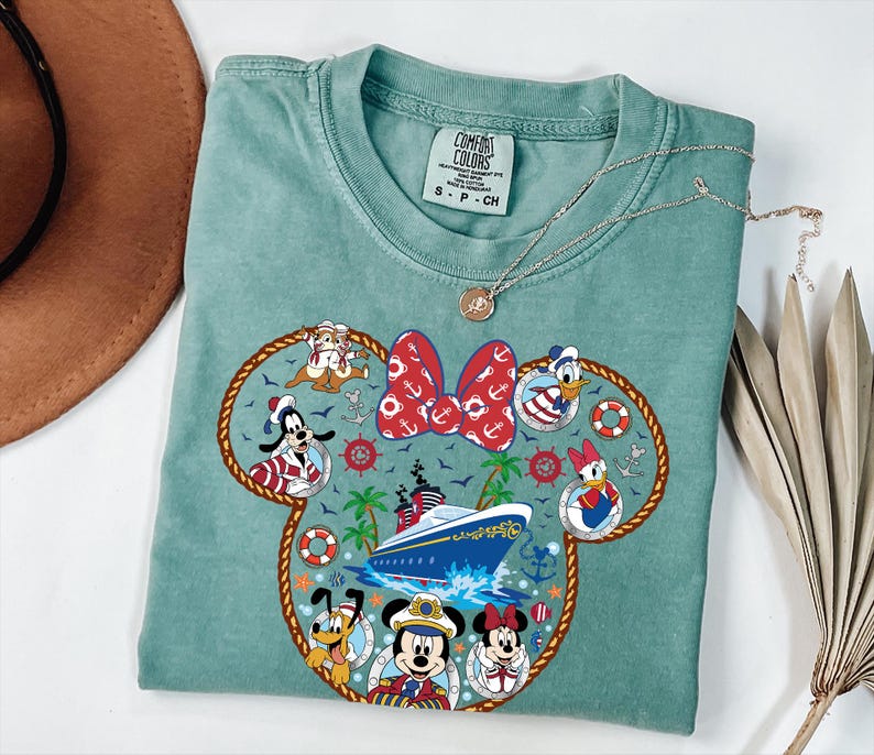 May include: A seafoam green t-shirt with a Minnie Mouse head design incorporating nautical themes. The design includes a red bow with white anchors, a cruise ship, and Disney characters in sailor outfits. A silver necklace is placed on the shirt.