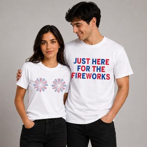 May include: Two white short-sleeved t-shirts. One shirt features two red, white, and blue firework graphics on the chest. The other shirt features the text "JUST HERE FOR THE FIREWORKS" in red and blue block lettering on the chest.