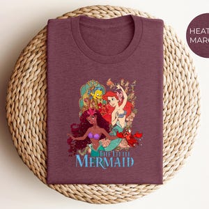 May include: Heather maroon t-shirt featuring a colorful graphic of mermaids and sea creatures. The design includes the text "THE LITTLE MERMAID" in blue. The shirt is displayed on a woven surface.