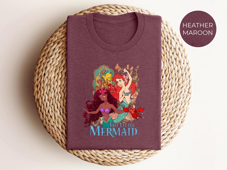 May include: Heather maroon t-shirt featuring a colorful graphic of mermaids and sea creatures. The design includes the text "THE LITTLE MERMAID" in blue. The shirt is displayed on a woven surface.