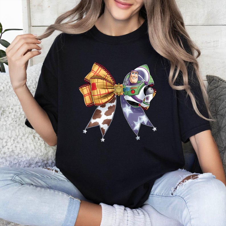 May include: Black t-shirt with a colourful bow design featuring a cartoon character. The bow incorporates a plaid pattern, a cow print, and a star-studded ribbon. The cartoon character is in a spacesuit.