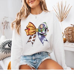 May include: White sweatshirt with a large bow design incorporating a Buzz Lightyear graphic. The bow features sections of plaid, cow print, and purple with star accents. The graphic is a colourful, cartoon-style design.