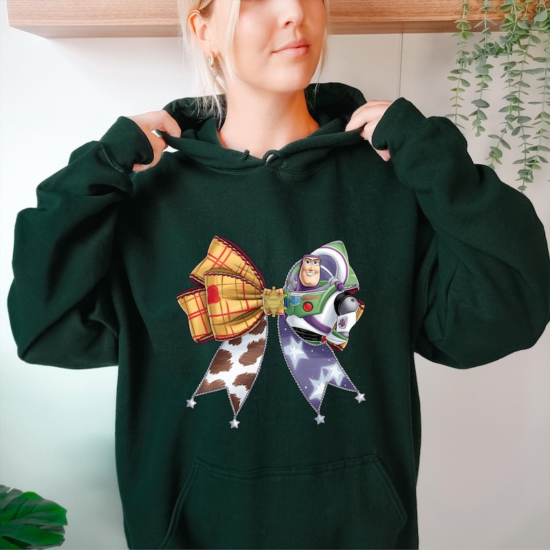 May include: Dark green hoodie featuring a colorful bow design with a Buzz Lightyear graphic. The bow has a plaid pattern, cow print, and a purple ribbon with white stars. The hoodie has a front pocket and a drawstring hood.