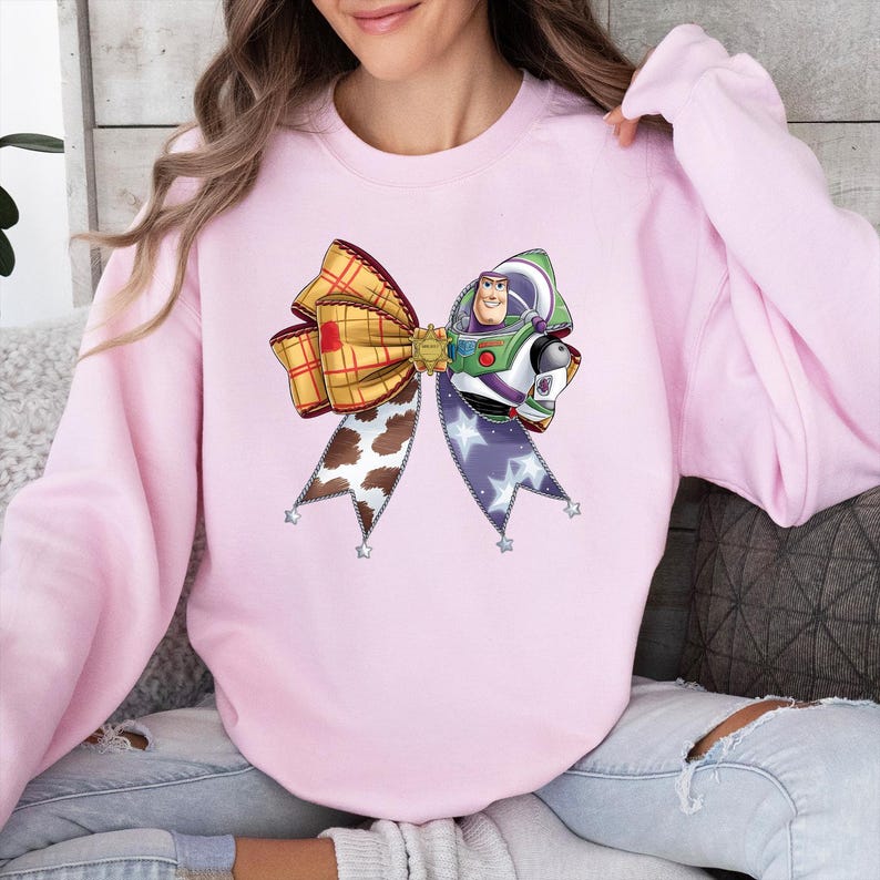 May include: A pale pink sweatshirt with a large bow design. The bow incorporates a plaid pattern, a cow print, and a star-patterned ribbon. The bow's centrepiece features a cartoon image of Buzz Lightyear.