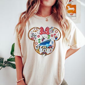 May include: Cream-coloured t-shirt with a Minnie Mouse-shaped design and a nautical theme. The design includes a red bow, a cruise ship, and Disney characters such as Mickey, Donald, and Goofy. The shirt has short sleeves.