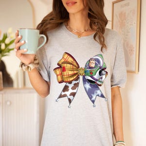 May include: A grey t-shirt with a large bow design. The bow incorporates a plaid pattern, a cowhide print, and a Buzz Lightyear character. The bow is decorated with star-shaped accents. The shirt is a casual wear item.