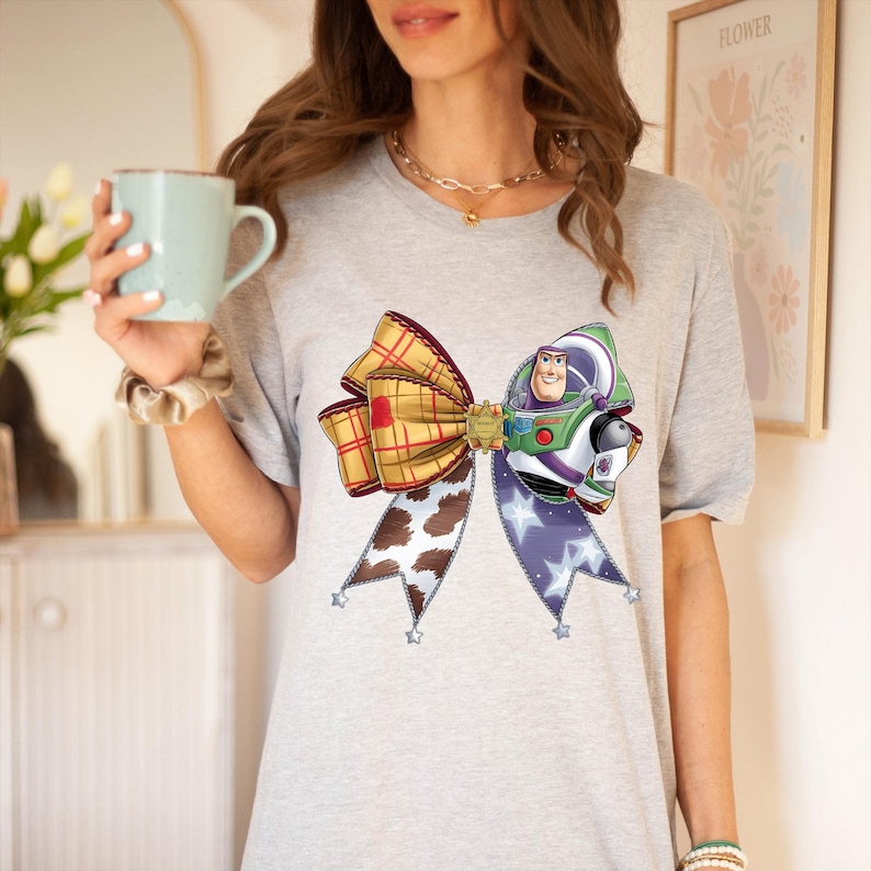 May include: A grey t-shirt with a large bow design. The bow incorporates a plaid pattern, a cowhide print, and a Buzz Lightyear character. The bow is decorated with star-shaped accents. The shirt is a casual wear item.