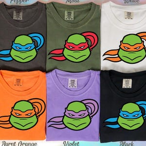 May include: Six t-shirts in various colors: pepper, moss, white, burnt orange, violet, and black. Each shirt features a cartoon turtle face with a colored mask and matching head wrap. The shirts are labeled with their respective color names.