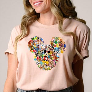 May include: A light pink t-shirt featuring a large Mickey Mouse head design composed of various Disney characters. The design includes characters from classic and modern Disney films, creating a colorful collage.