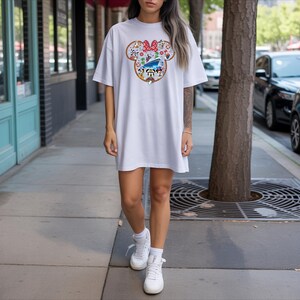 May include: White t-shirt dress with a colourful Minnie Mouse graphic. The graphic features a red bow, flowers, and Disney characters. The dress is worn with white trainers and socks. The person is standing on a pavement next to a tree.