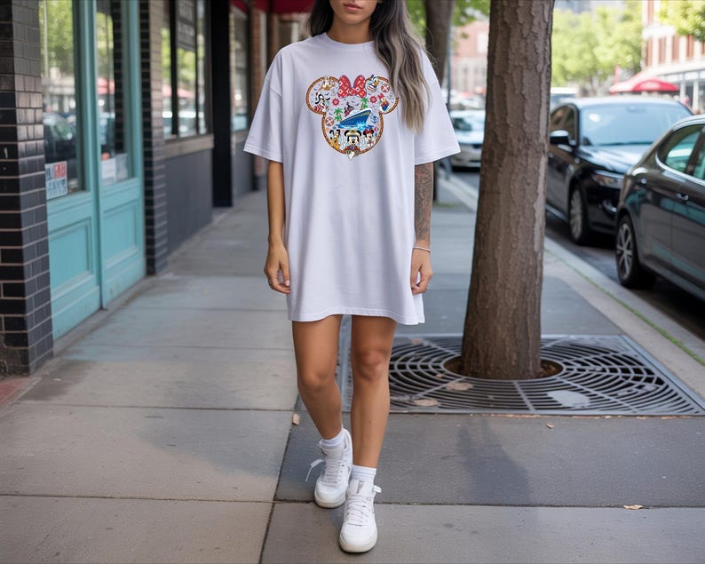 May include: White t-shirt dress with a colourful Minnie Mouse graphic. The graphic features a red bow, flowers, and Disney characters. The dress is worn with white trainers and socks. The person is standing on a pavement next to a tree.