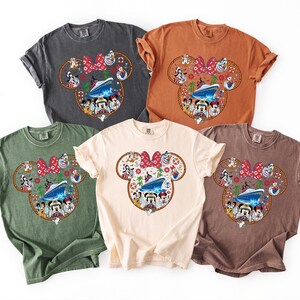 May include: Five short-sleeved t-shirts in shades of grey, orange, green, cream, and brown. Each shirt has a Mickey Mouse head design with a cruise ship, characters, and a red bow. The shirts are in various colours.