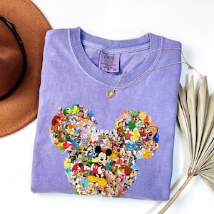 May include: A light purple Comfort Colors t-shirt featuring a Mickey Mouse head design filled with various Disney characters. The shirt has a crew neck and short sleeves. A gold necklace with a pendant is draped on the shirt.