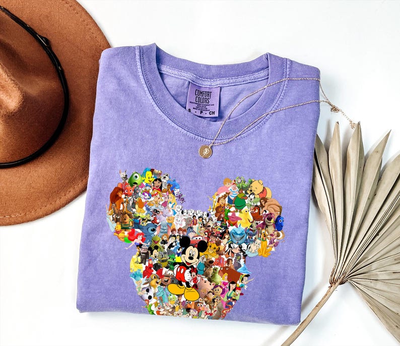 May include: A light purple Comfort Colors t-shirt featuring a Mickey Mouse head design filled with various Disney characters. The shirt has a crew neck and short sleeves. A gold necklace with a pendant is draped on the shirt.