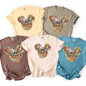 May include: Five short-sleeved t-shirts in various colors: yellow, grey, brown, cream, and blue. Each shirt features a Mickey Mouse head design made up of various cartoon characters. The shirts are made of a soft material.