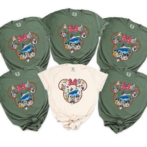 May include: Six t-shirts in shades of olive green and cream. Each shirt displays a Mickey Mouse head design with a cruise ship, characters, and a red bow. The shirts are short-sleeved and likely made of cotton.