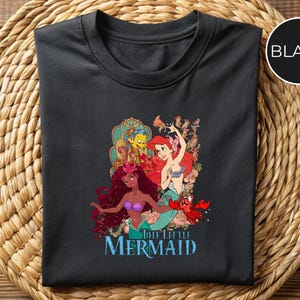 May include: Black t-shirt featuring a colorful graphic of characters from "The Little Mermaid." The design includes Ariel, Flounder, Sebastian, and other mermaids. The words "The Little Mermaid" are printed in blue below the graphic.
