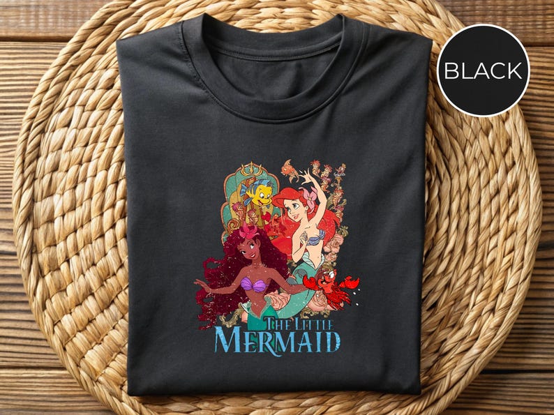 May include: Black t-shirt featuring a colorful graphic of characters from "The Little Mermaid." The design includes Ariel, Flounder, Sebastian, and other mermaids. The words "The Little Mermaid" are printed in blue below the graphic.