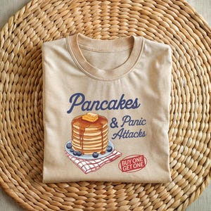 May include: A beige t-shirt with a graphic of a stack of pancakes with syrup and butter, and the text "Pancakes & Panic Attacks" in blue. The text "Buy One Get One" is in red.