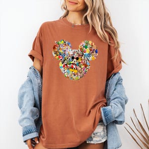 May include: A rust-colored t-shirt featuring a Mickey Mouse head design composed of various Disney characters. The graphic is centered on the shirt, showcasing a collage of colorful cartoon figures. The shirt has short sleeves and a relaxed fit.