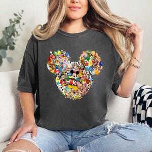May include: Dark gray t-shirt featuring a Mickey Mouse head design composed of various Disney characters. The graphic includes characters like Mickey, Minnie, Donald, and Goofy, among others. The shirt is a short-sleeved, casual style.