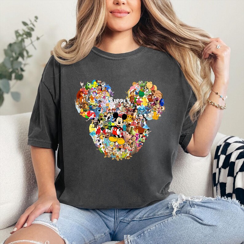 May include: Dark gray t-shirt featuring a Mickey Mouse head design composed of various Disney characters. The graphic includes characters like Mickey, Minnie, Donald, and Goofy, among others. The shirt is a short-sleeved, casual style.
