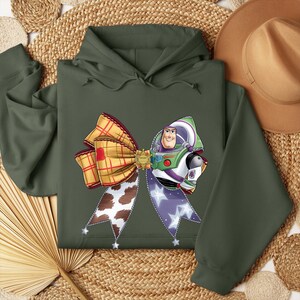 May include: An olive green hoodie with a large bow design incorporating elements from the film Toy Story. The bow features a plaid pattern, a cow print, and a Buzz Lightyear graphic. The hoodie has a drawstring hood and long sleeves.