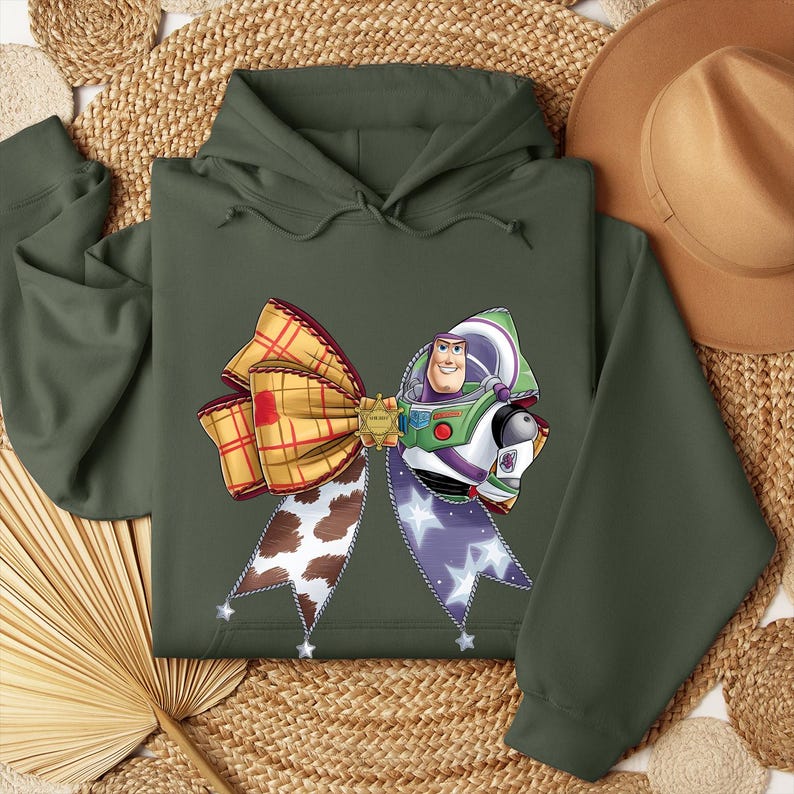 May include: An olive green hoodie with a large bow design incorporating elements from the film Toy Story. The bow features a plaid pattern, a cow print, and a Buzz Lightyear graphic. The hoodie has a drawstring hood and long sleeves.