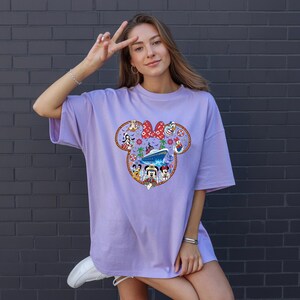 May include: A lilac t-shirt with a colourful Minnie Mouse graphic. The design incorporates Minnie's bow, Disney characters, and a cruise ship. The t-shirt has a loose fit.
