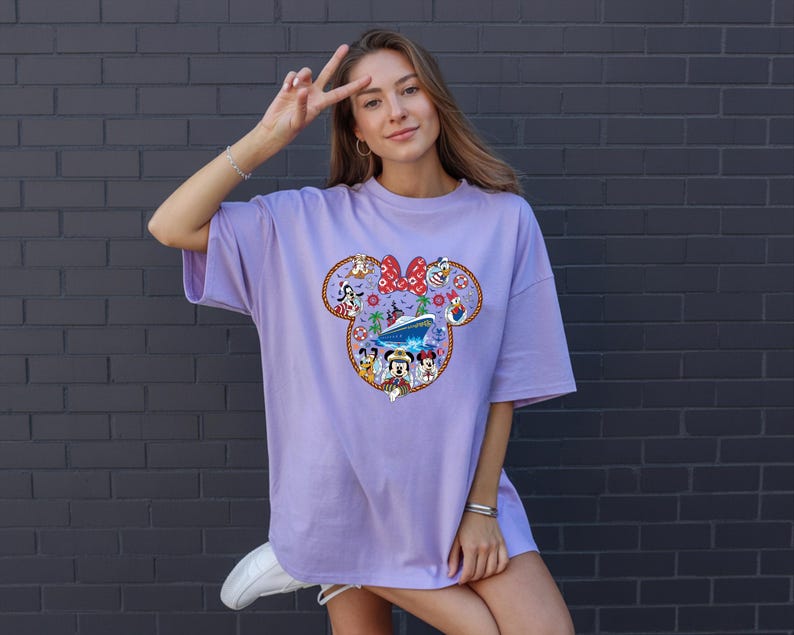 May include: A lilac t-shirt with a colourful Minnie Mouse graphic. The design incorporates Minnie's bow, Disney characters, and a cruise ship. The t-shirt has a loose fit.