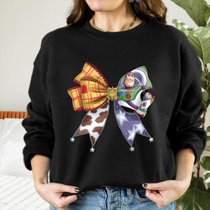 May include: Black sweatshirt with a large, colourful bow design. The bow incorporates elements of a plaid pattern, cow print, and a Buzz Lightyear character. The bow is adorned with star-shaped accents.