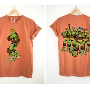 May include: Terracotta-colored t-shirt featuring a graphic of a Teenage Mutant Ninja Turtle on the front and the group on the back. The front shows a single turtle, while the back displays all four turtles. The shirt has rolled-up sleeves.