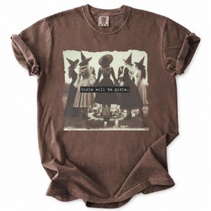 May include: A brown short-sleeved t-shirt featuring a central graphic print of six figures wearing pointed hats and dresses standing in a circle. The text "Girls will be girls." is printed in a black rectangular box across the center of the image.