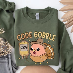 May include: An olive green sweatshirt with the text "CODE GOBBLE" in a playful font. A cartoon turkey wearing a hat and a gravy IV bag is featured. The design is perfect for Thanksgiving or a fun fall outfit.