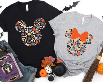 May include: Two t-shirts with Halloween-themed designs. One black t-shirt features a Mickey Mouse head made of Halloween icons. The other is gray with an orange bow and a similar design. Halloween decorations are scattered around.