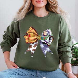 May include: An olive green sweatshirt with a large bow design featuring a Buzz Lightyear graphic. The bow incorporates a plaid pattern, cow print, and a purple ribbon adorned with white stars. The Buzz Lightyear graphic is centrally positioned.