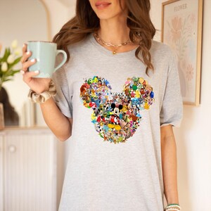 May include: A light gray t-shirt featuring a colorful collage of Disney characters arranged in the shape of Mickey Mouse's head. The design includes Mickey Mouse in the center, surrounded by other characters. The shirt has short sleeves.