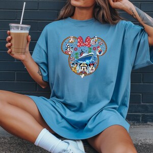 May include: A pale blue t-shirt with a Mickey Mouse head design filled with Disney characters and a cruise ship. The design includes a red bow, various characters, and a nautical theme. The shirt is worn by a person.