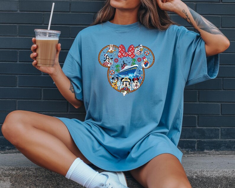 May include: A pale blue t-shirt with a Mickey Mouse head design filled with Disney characters and a cruise ship. The design includes a red bow, various characters, and a nautical theme. The shirt is worn by a person.