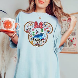 May include: Pale blue t-shirt with a Minnie Mouse-shaped design incorporating nautical themes. The design includes a cruise ship, Mickey Mouse, Minnie Mouse, Donald Duck, and Goofy. The shirt is worn with a white mug featuring an orange sun.