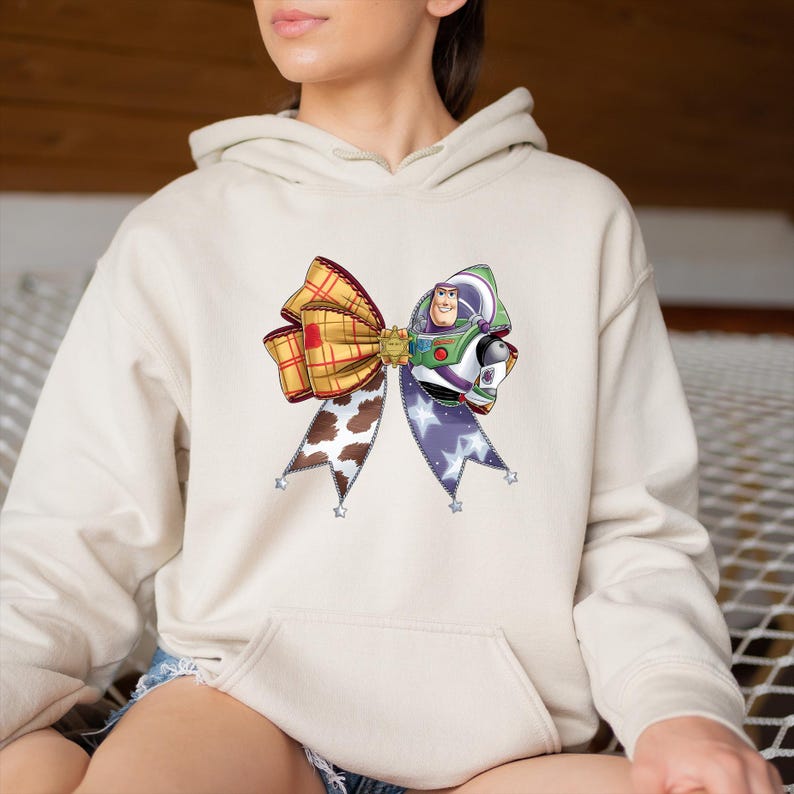 May include: A beige hoodie featuring a colorful bow design. The bow is composed of a plaid section, a cow print section, and a section with a purple star pattern. The bow also features a graphic of Buzz Lightyear.