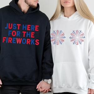 May include: A black hooded sweatshirt with the text JUST HERE FOR THE FIREWORKS in red and blue, and a white hooded sweatshirt featuring two circular red and blue firework burst graphics on the chest, both with front kangaroo pockets.