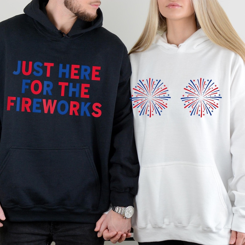 May include: A black hooded sweatshirt with the text JUST HERE FOR THE FIREWORKS in red and blue, and a white hooded sweatshirt featuring two circular red and blue firework burst graphics on the chest, both with front kangaroo pockets.