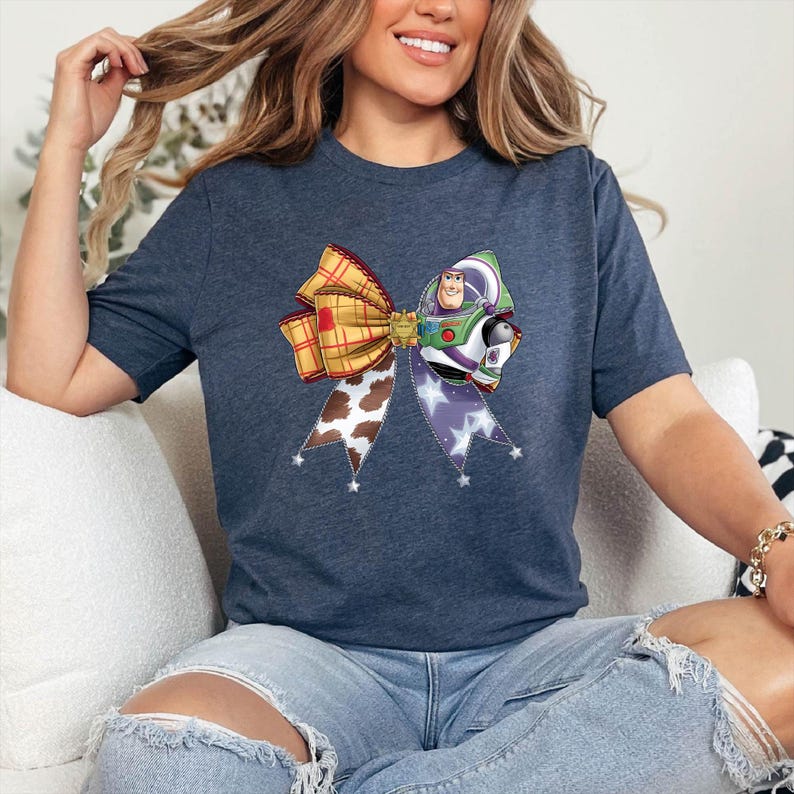 May include: Navy blue t-shirt with a large bow design. The bow incorporates plaid, cow print, and a Buzz Lightyear graphic. The bow is stitched with a purple star pattern.