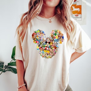 May include: Cream-colored t-shirt featuring a large Mickey Mouse head design. The head is composed of a collage of various Disney characters in vibrant colors. The shirt has short sleeves and a relaxed fit, suitable for casual wear.