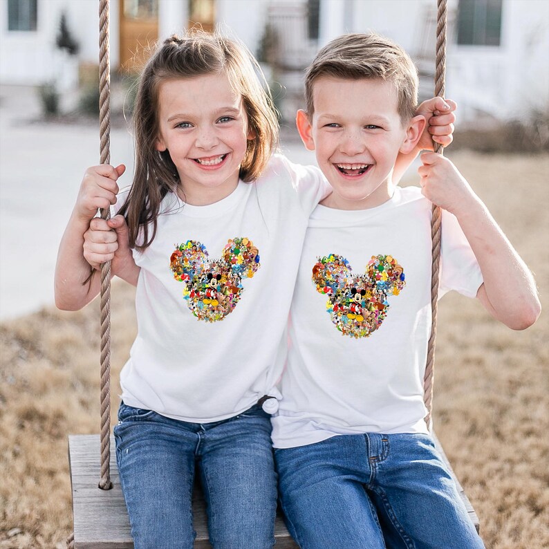 May include: Two children wearing white t-shirts with a colorful Mickey Mouse head design made of various cartoon characters. They are sitting on a wooden swing, smiling, and wearing blue jeans.