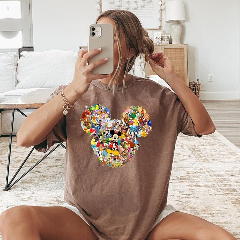 May include: Brown t-shirt featuring a Mickey Mouse head design composed of various Disney characters. The shirt is a neutral color and has a relaxed fit. The design includes characters like Mickey, Minnie, Donald, and Goofy.
